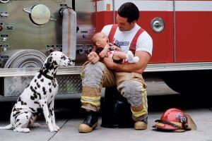 This is a photograph of a dalmatian sitting and watching a firefighter cradle an infant.